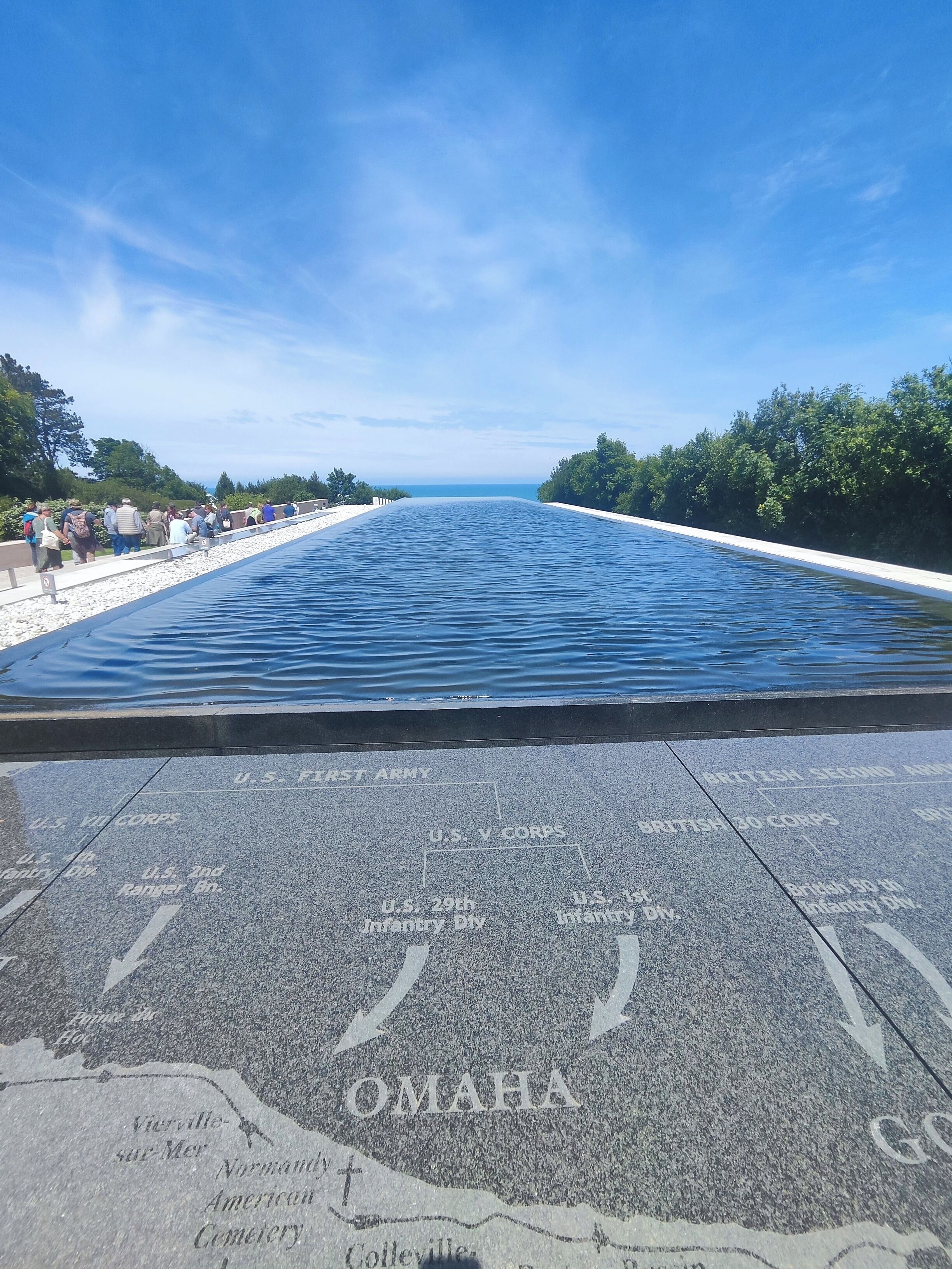 Una piscina larga y estrecha con un cielo azul despejado arriba. La piscina está rodeada por un camino empedrado con inscripciones de unidades militares y la palabra "OMAHA".