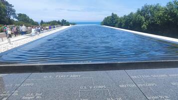 Una piscina larga y estrecha con un cielo azul despejado arriba. La piscina está rodeada por un camino empedrado con inscripciones de unidades militares y la palabra "OMAHA".