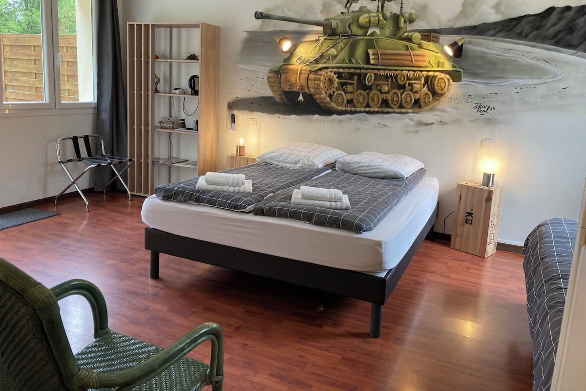 A bedroom with a bed, a chair, a wardrobe, and a wall mural of a tank.