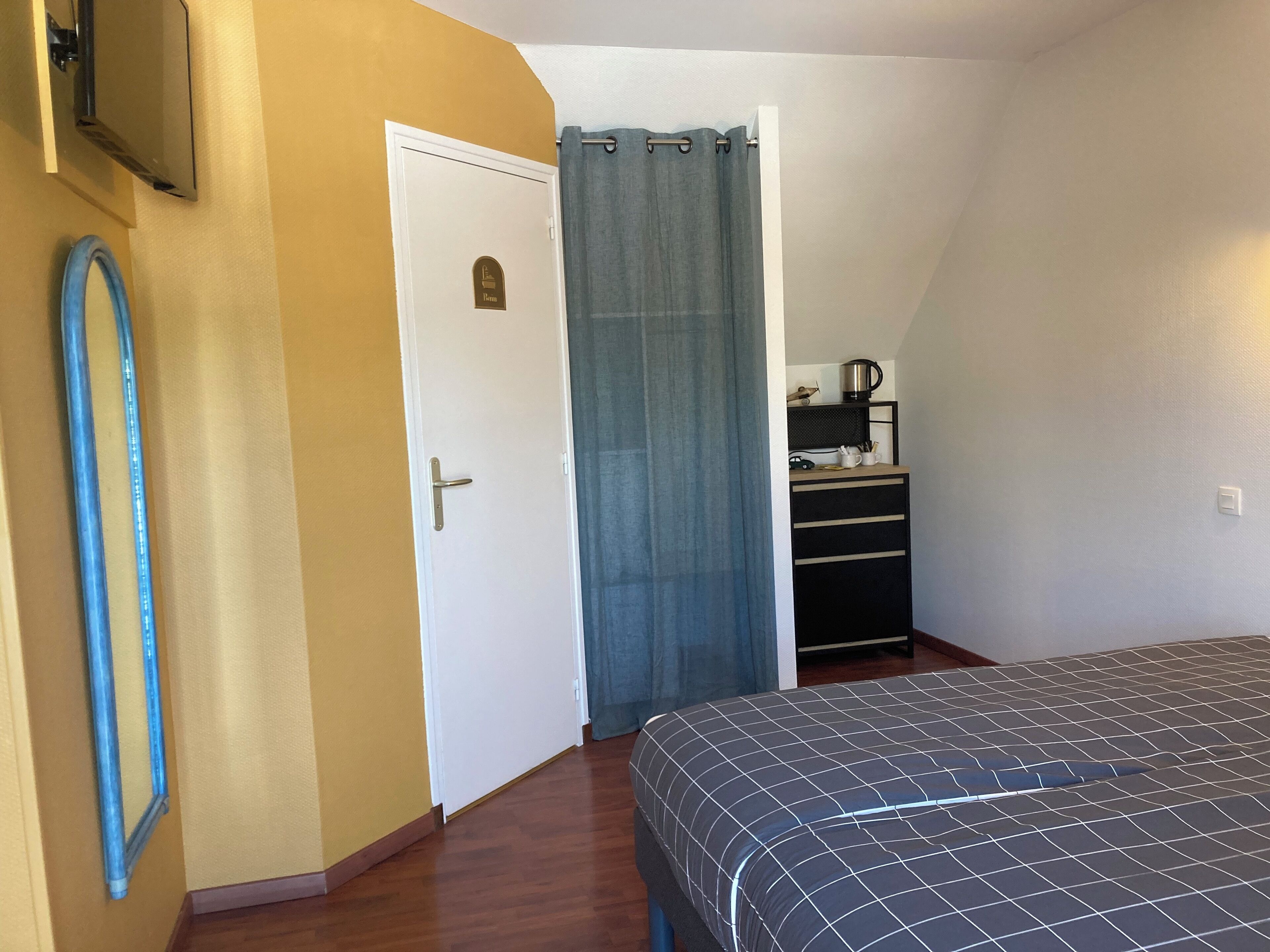 Une chambre à coucher avec un lit, une porte, un rideau, un petit bureau avec une bouilloire et un téléviseur fixé au mur.