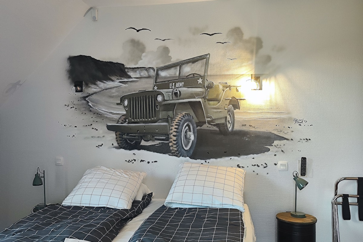 A bedroom with two beds, a wall mural of a military jeep, and a bedside lamp on a barrel.