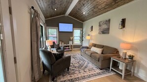 A cozy living room with a wooden ceiling, a large rug, a sofa, a chair, and a TV in the background. - The Crepe Myrtle Cottage (Franklin)