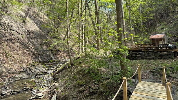 Eine Holzbrücke führt zu einer rustikalen Holzhütte in einem bewaldeten Gebiet mit einem Bach.