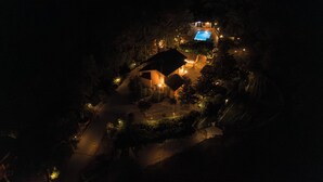 An aerial view of a property at night with a lit swimming pool, a house, and surrounding vegetation.
