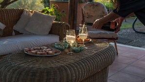A wicker table with plates of food and glasses of white wine, set outdoors with a wicker sofa and floral chair in the background.