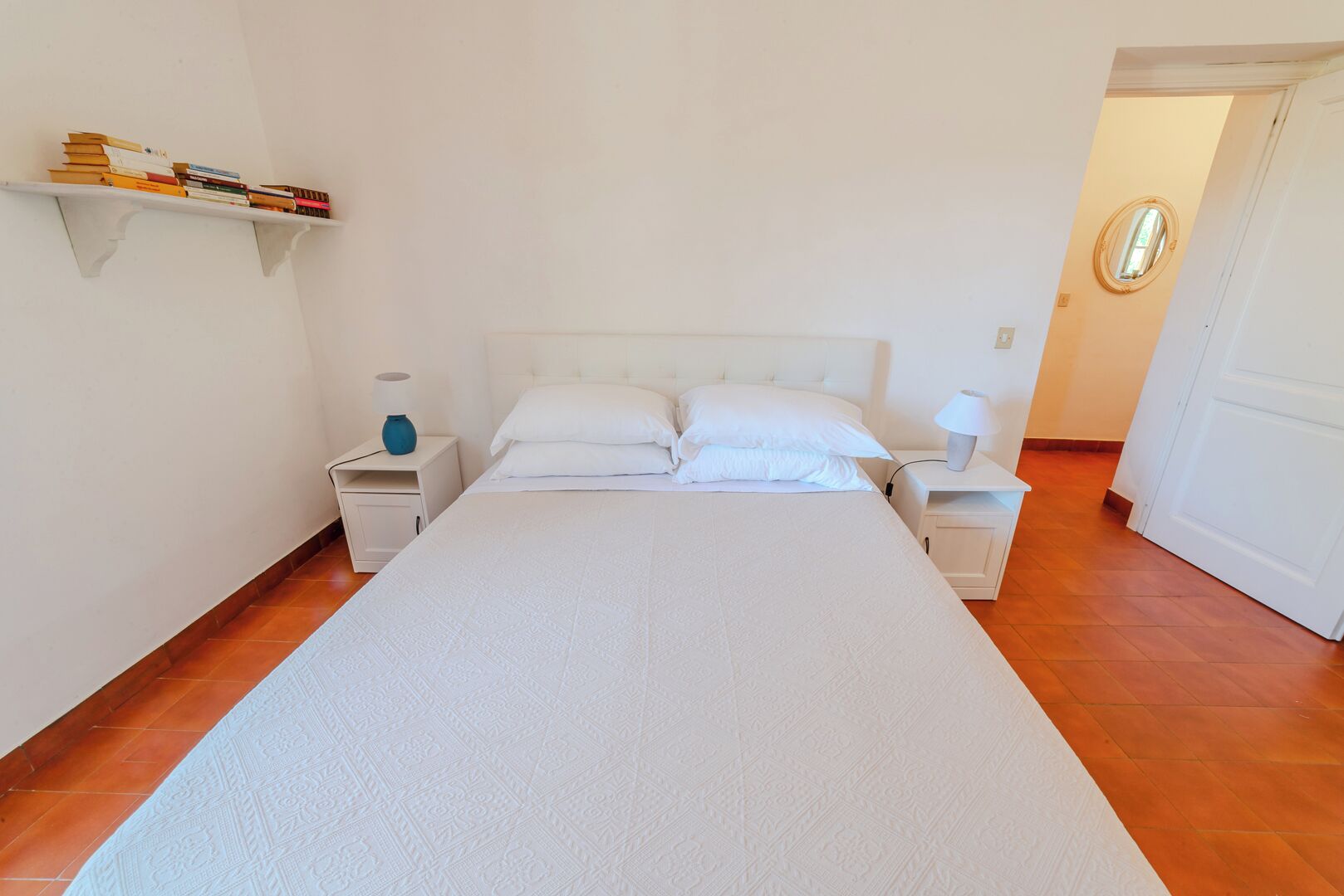 Une chambre à coucher avec un grand lit, deux tables de chevet, une étagère remplie de livres et une porte donnant sur une autre pièce.