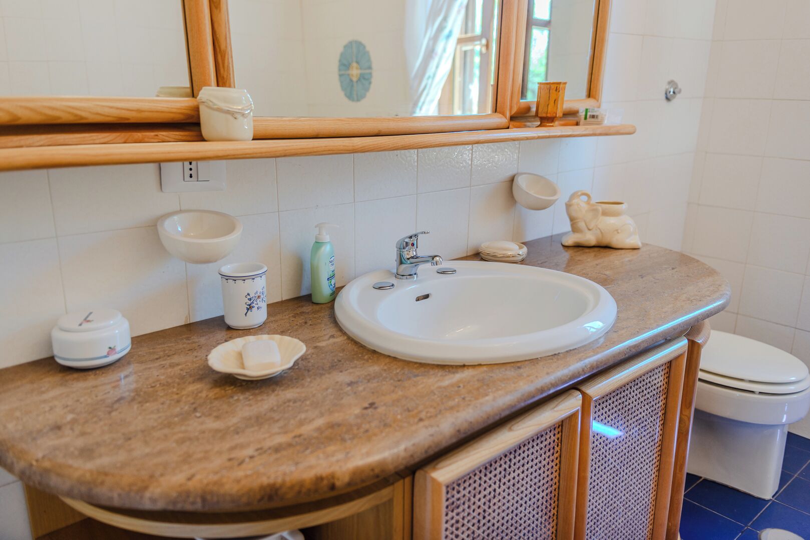 Un baño con lavamanos blanco, espejo, repisa de madera con artículos de aseo y un inodoro.