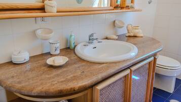 Un baño con lavamanos blanco, espejo, repisa de madera con artículos de aseo y un inodoro.