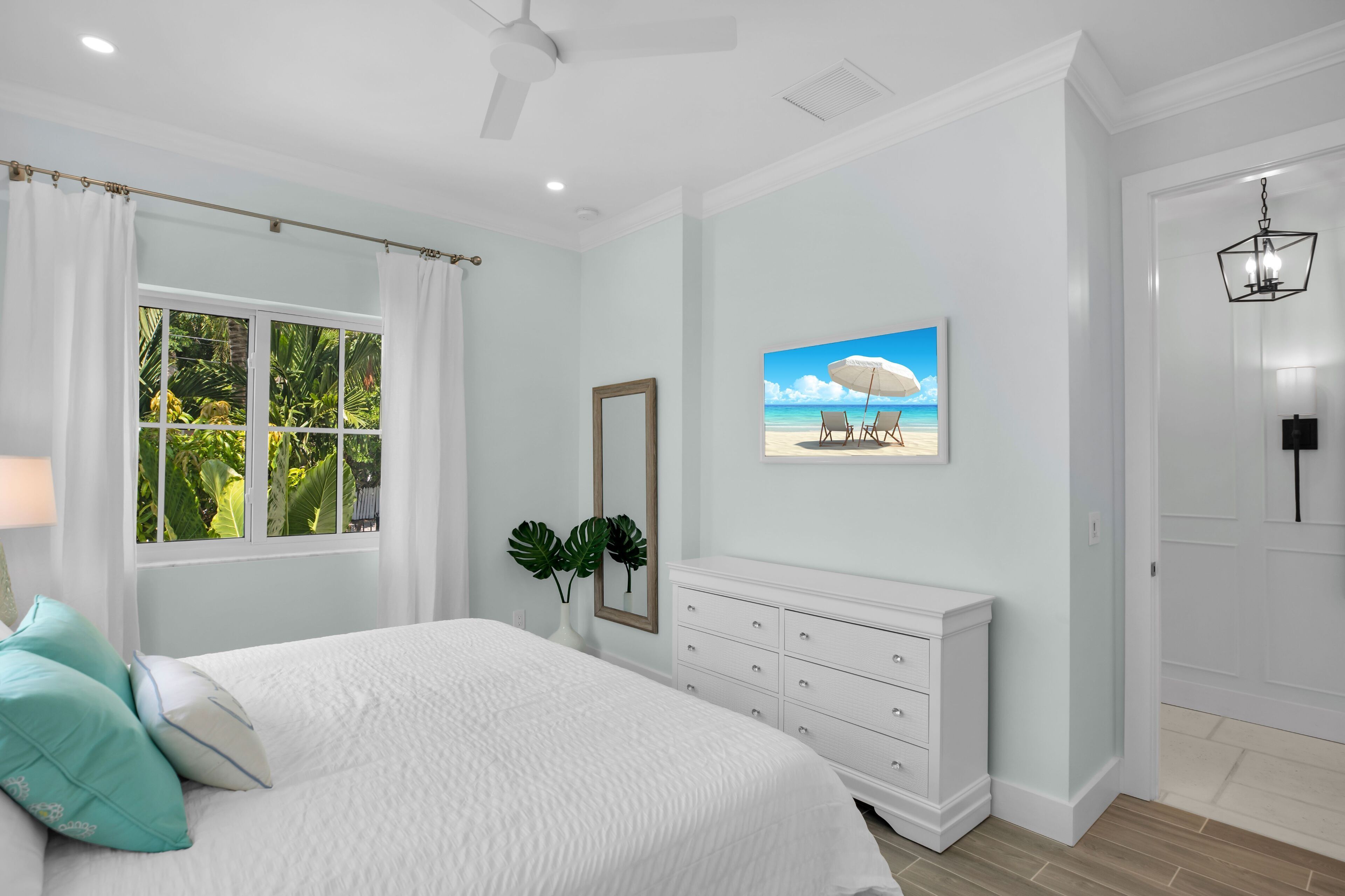 Une chambre à coucher comprenant un lit, une commode, une fenêtre avec des rideaux, un ventilateur de plafond, un miroir fixé au mur et un tableau sur le thème de la plage.