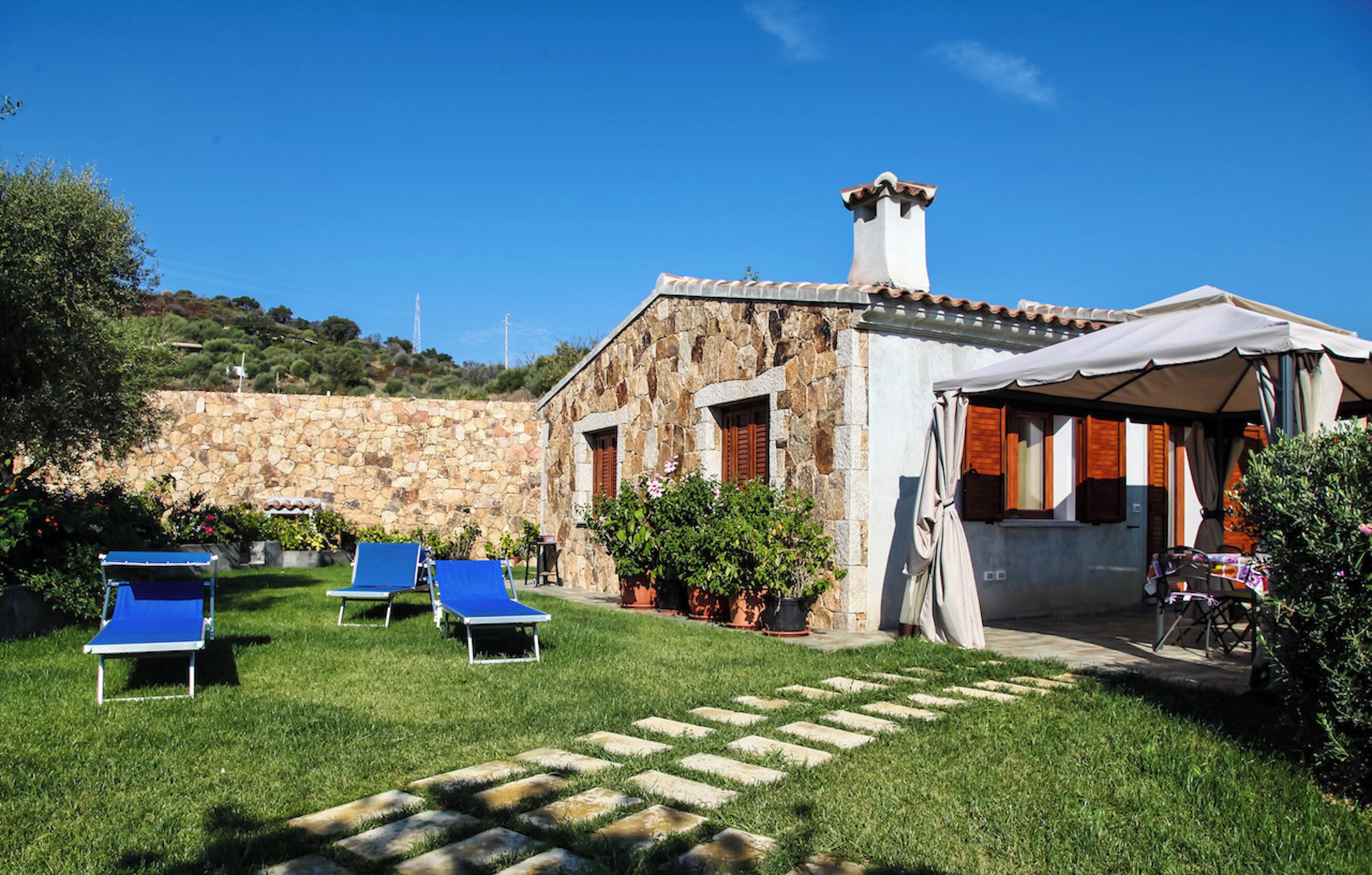 A stone building with a tiled roof, a covered outdoor seating area, and a grassy lawn with blue lounge chairs.