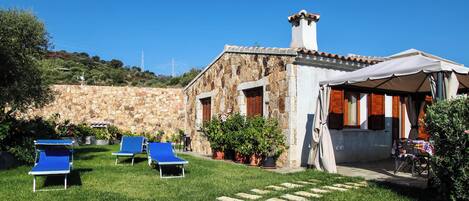 A stone building with a tiled roof, a covered outdoor seating area, and a grassy lawn with blue lounge chairs.