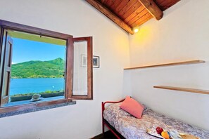 Una habitación pequeña con cama, ventana con vista a un lago y colinas, y techo de vigas de madera.