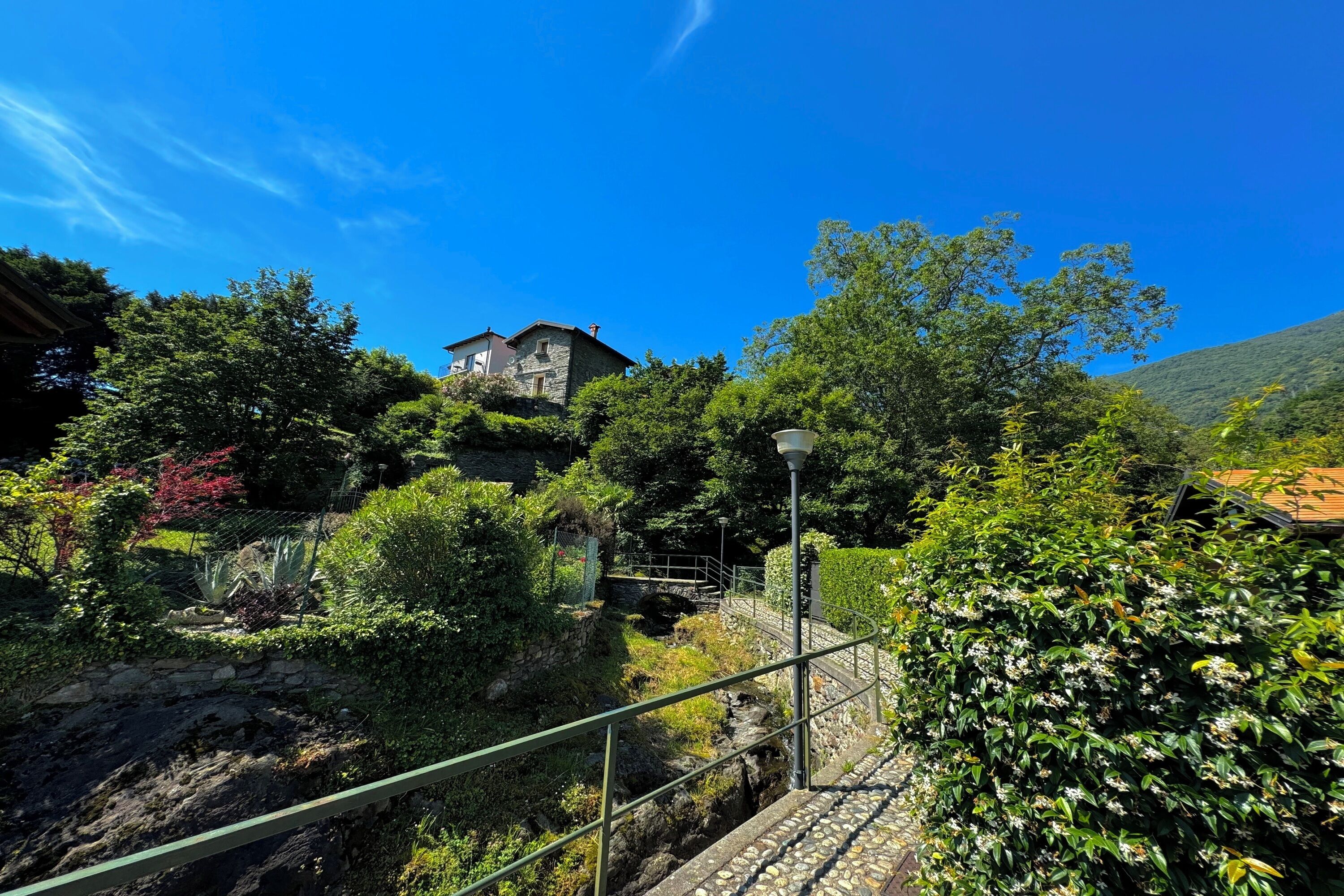 A stone house with a stone staircase leading up to it, surrounded by lush greenery and a clear blue sky.