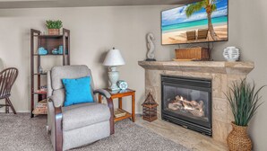 A living room with a fireplace, a comfortable armchair, a bookshelf, and a TV displaying a beach scene. - Top Floor, Ocean View Condo in The Heart of The Taft District in Lincoln City (Lincoln City)
