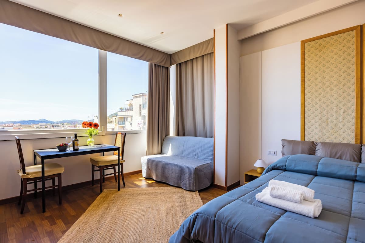 A hotel room with a large window, a desk with a chair, a sofa, a bed with a blue bedspread, and a small nightstand with a lamp.