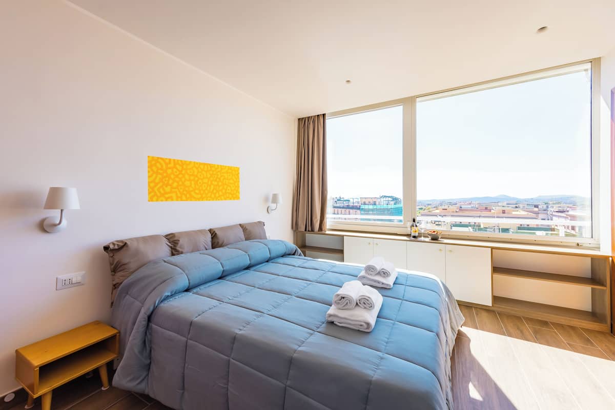 A modern hotel room with a large bed, a desk with a view, and a cityscape visible through the window.