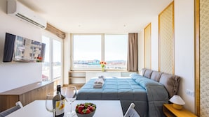 A hotel room with a bed, a dining table with wine glasses and a bowl of fruit, a TV on the wall, and a large window with a city view. - Chiave di Fuga (Naples)