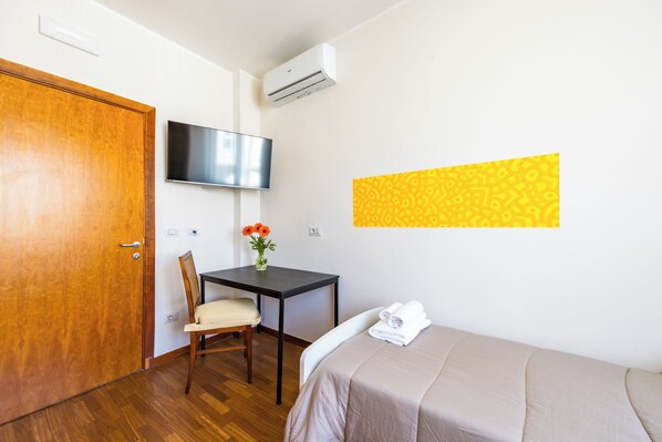 Een hotelkamer met een houten deur, een bureau met stoel, een bed met beige sprei, een flatscreen televisie aan de muur en een airconditioner.