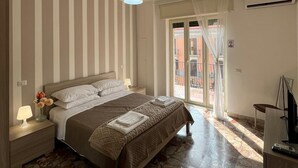 Une chambre d’hôtel comprenant un lit, une table de chevet, une lampe, un vase avec des fleurs, un luminaire au plafond, une porte-fenêtre coulissante donnant sur l’extérieur et un téléviseur posé sur un meuble.