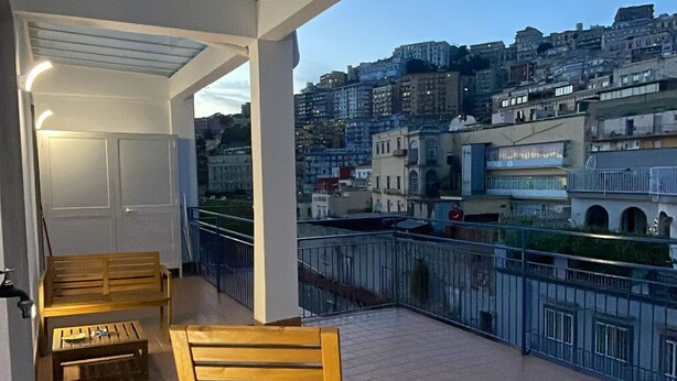 Ein Balkon mit einem Holzstuhl und einem kleinen Tisch, mit Blick auf eine Stadtlandschaft mit Gebäuden.