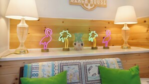 Une chambre avec une tête de lit en bois, deux lampes, un néon indiquant « BEACH HOUSE », et un oreiller matelassé sur le lit.