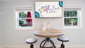 Une pièce avec une table ronde, deux tabourets, un téléviseur affichant le logo de « The Best Rentals », et deux fenêtres donnant sur un paysage où l’on aperçoit un parasol rouge et de la verdure.