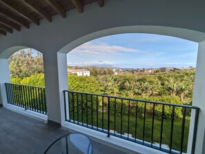 A balcony with a glass table and a view of a lush garden and distant buildings.