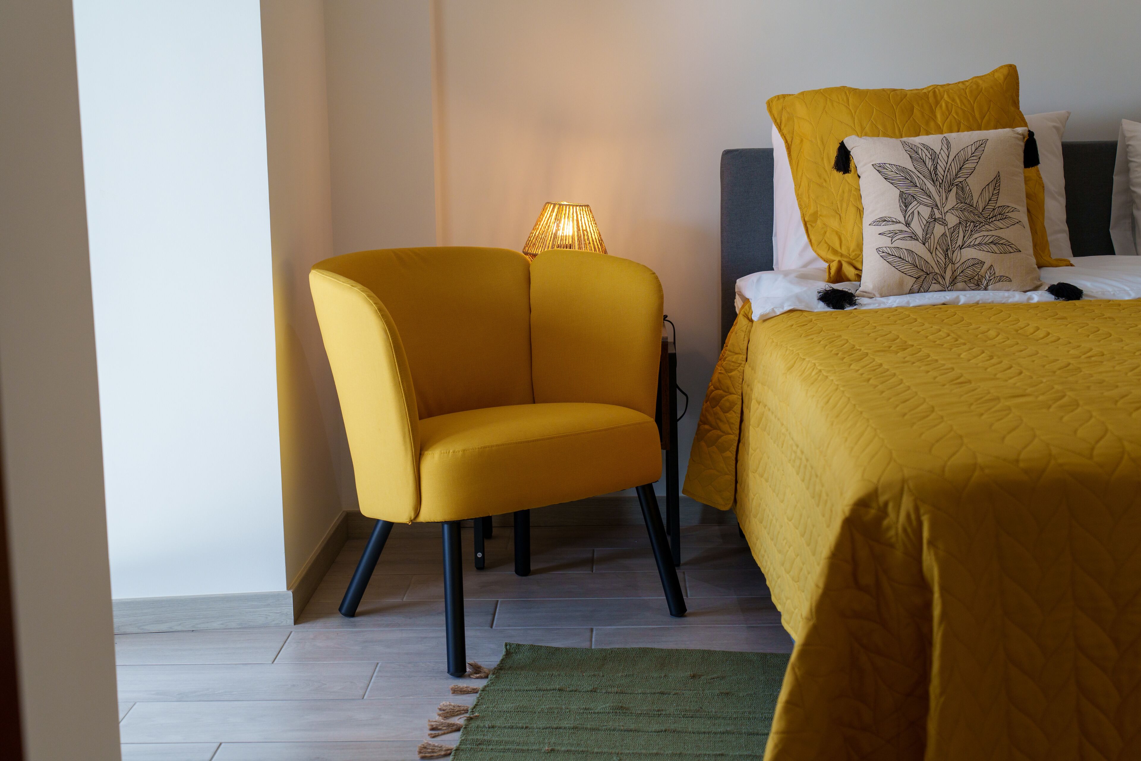 A room with a bed, a yellow armchair, a lamp, and a green rug.