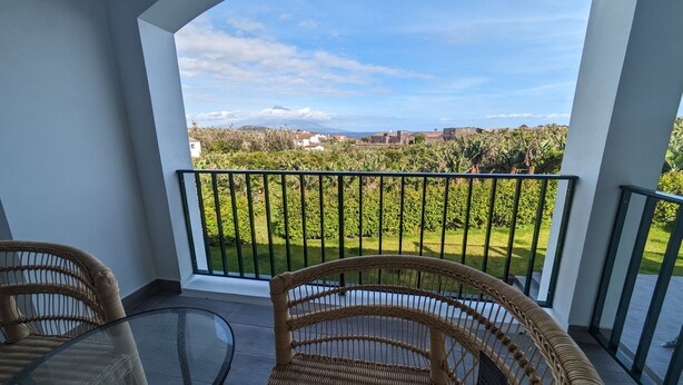 Varanda com mobiliário de vime e mesa de vidro, com vista para um jardim exuberante e edifícios ao longe.