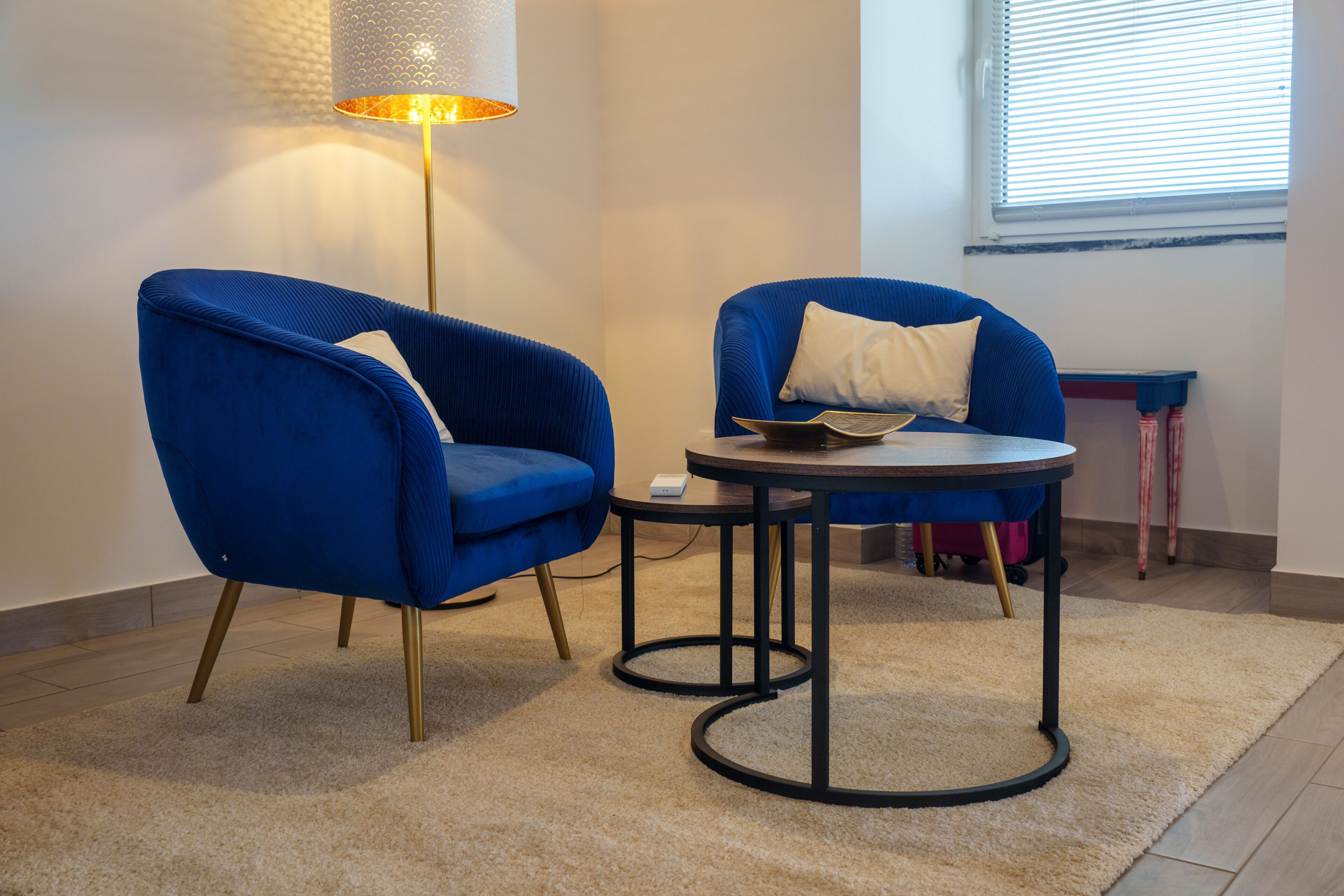 A room with two blue armchairs, a small round table, a lamp, and a window with blinds.