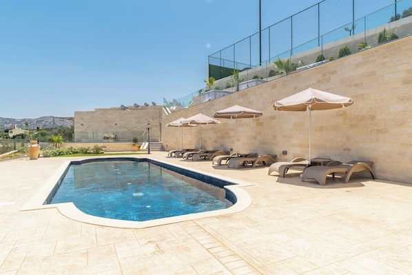 Piscina con sillas de descanso y sombrillas, rodeada por un muro de piedra y una barandilla de vidrio.