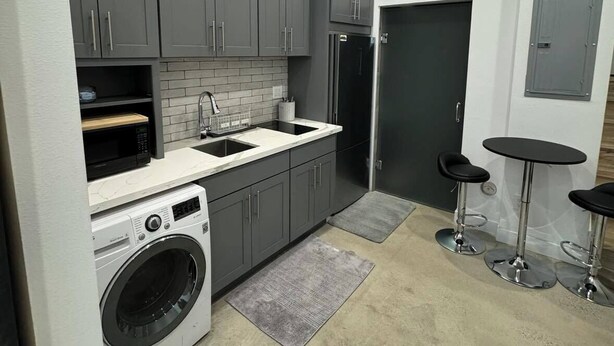 A compact kitchen with grey cabinets, a built-in washing machine, a microwave, and a refrigerator. There are two bar stools and a small table. - Cozy studio in enchanting Seattle with AC, WiFi (Seattle)