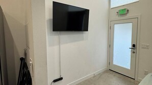 A room with a flat-screen TV mounted on the wall, a door with an exit sign, and a window with blinds. - Cozy studio in enchanting Seattle with AC, WiFi (Seattle)