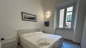 Een slaapkamer met een bed, een nachtkastje, een lampje aan de muur, een raam met uitzicht en een ingelijste foto.