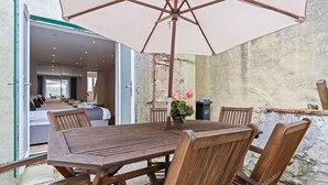 Eine offene Terrasse mit einem Holztisch und Stühlen, einem großen Sonnenschirm und einem Blumentopf mit rosa Blüten.