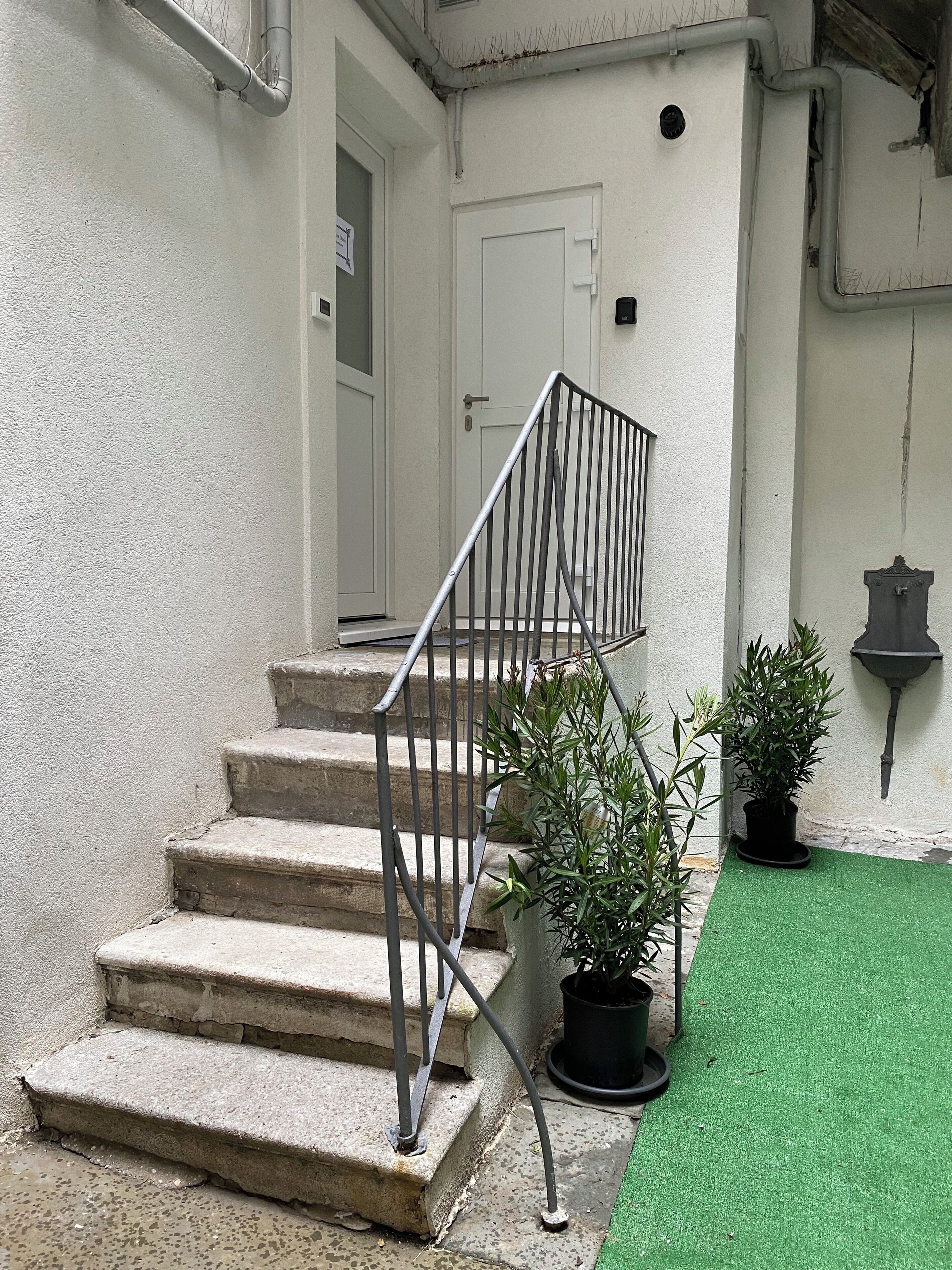 Een betonnen trap met metalen leuningen leidt naar een witte deur. Op de begane grond staan twee plantenpotten.