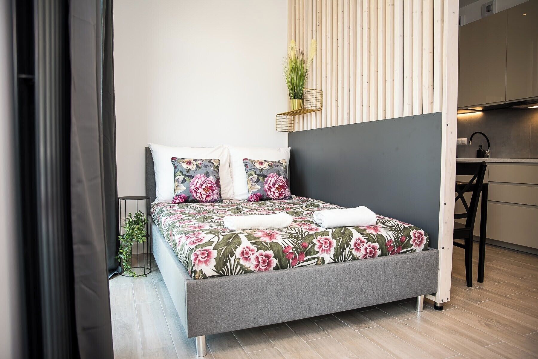 Un dormitorio moderno con una cama gris, ropa de cama floral, una pequeña planta en maceta y una cocina visible a la derecha.