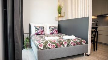 Un dormitorio moderno con una cama gris, ropa de cama floral, una pequeña planta en maceta y una cocina visible a la derecha.
