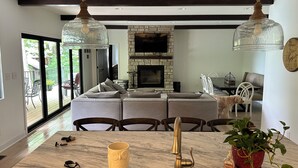 A modern living room with a stone fireplace, a dining area with a marble table, and a large window overlooking a deck.