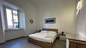A bedroom with a wooden bed, a nightstand, a wall-mounted picture, and a window with blinds. - Faro Porto Apartment 4 (Monte Argentario)