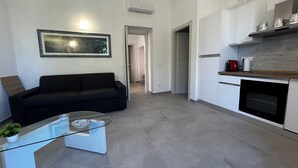 A modern living room with a black sofa, a glass coffee table, and a framed picture on the wall. A kitchen with white cabinets and a red toaster is visible. - Faro Porto Apartment 4 (Monte Argentario)