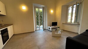 A modern living room with a flat-screen TV, a glass coffee table, and a black sofa. There is a kitchen area with a stove and oven, and a sliding glass door leading to a balcony. - Faro Porto Apartment 4 (Monte Argentario)