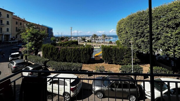 A balcony with a view of a marina, parked cars, and buildings. - Faro Porto Apartment 4 (Monte Argentario)