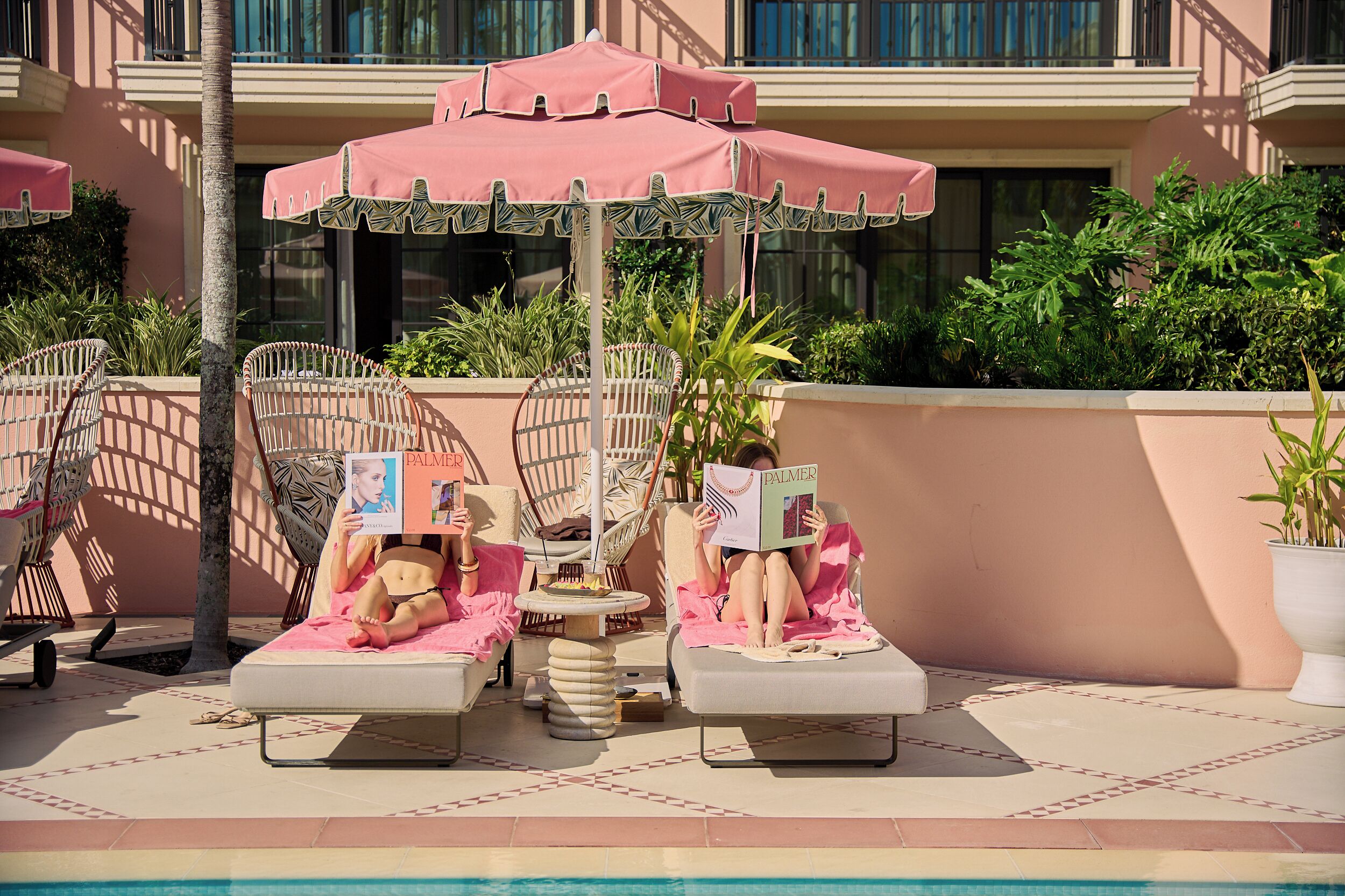 A poolside area with lounge chairs and umbrellas, a palm tree, and a building in the background.