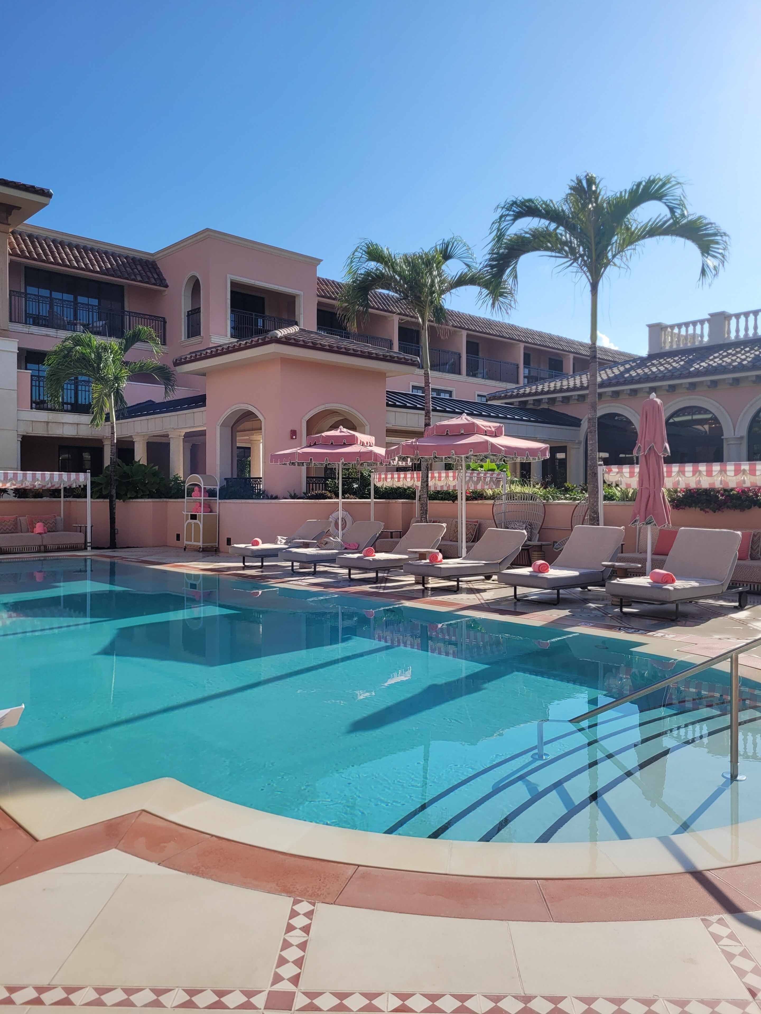Ein Swimmingpool mit Liegestühlen und Sonnenschirmen, umgeben von einem Gebäude mit Balkonen und Palmen.
