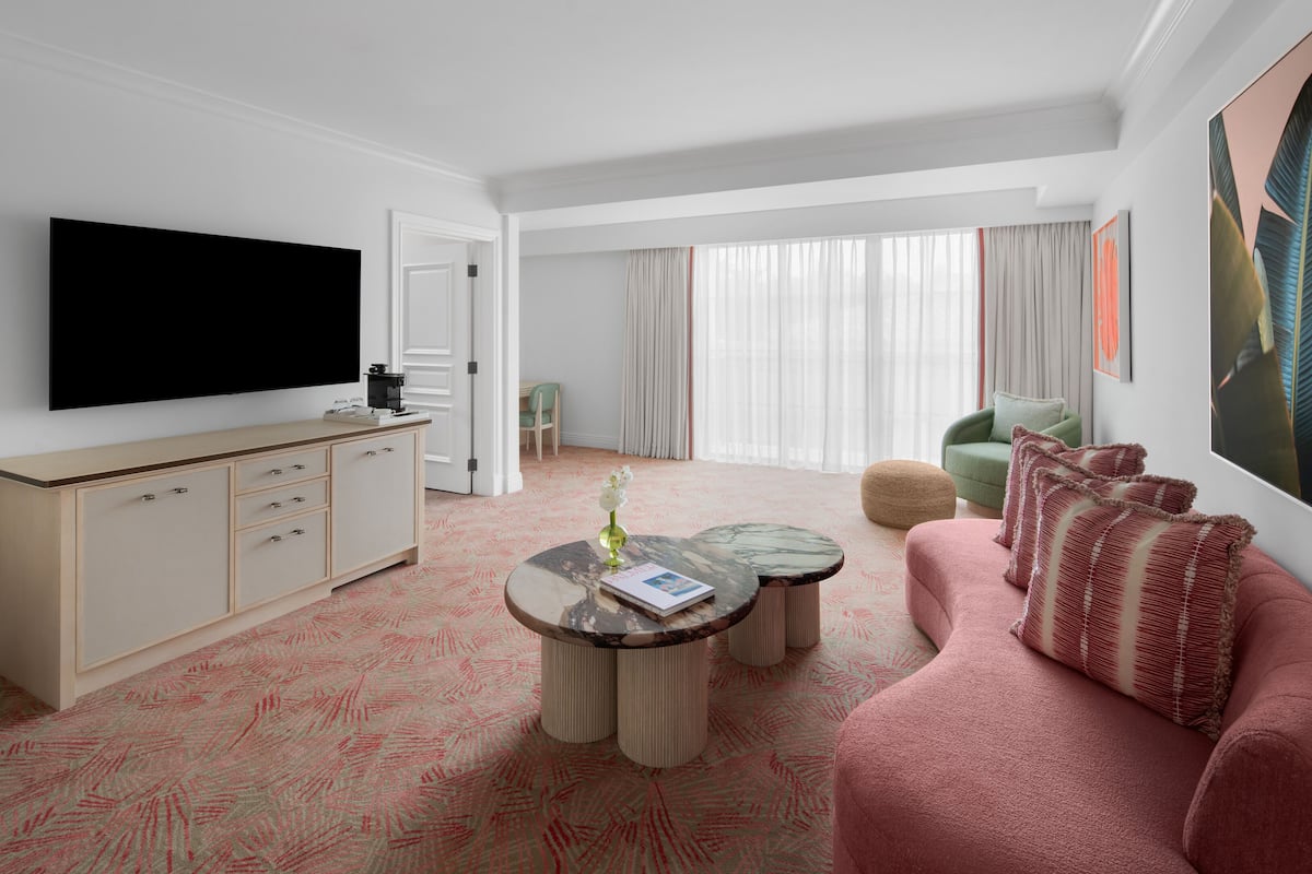 A modern hotel room with a flat-screen TV, a beige sofa, a pink couch, and a dining area with a table and chairs.
