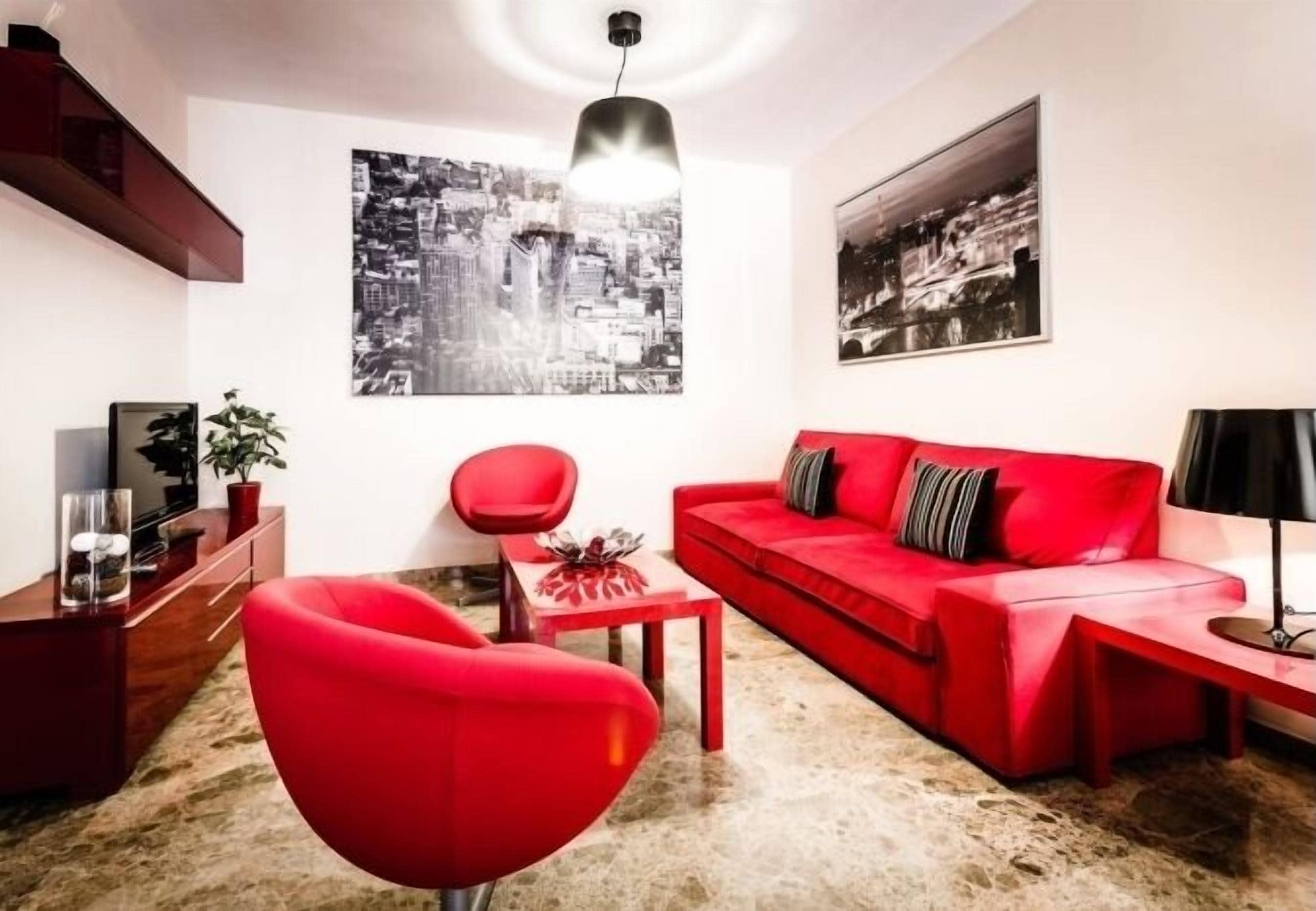 Een moderne woonkamer met een rode bank, een rode fauteuil en een salontafel. Op de muren hangen twee zwart-wit foto’s van een stadsgezicht.