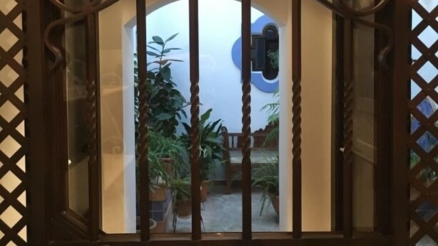 Una reja de hierro forjado con detalles decorativos en la parte superior, que da acceso a un patio con plantas en macetas y un ventanal azul circular.