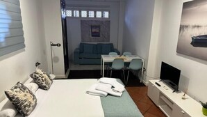 Habitación de hotel pequeña con una cama, un escritorio, un televisor y vista a un sofá azul y un cuadro al fondo.