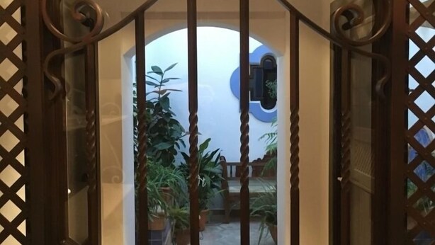 Un cancello in ferro battuto con un motivo a reticolo decorativo, che dà accesso a uno spazio esterno con piante in vaso e una finestra circolare blu.